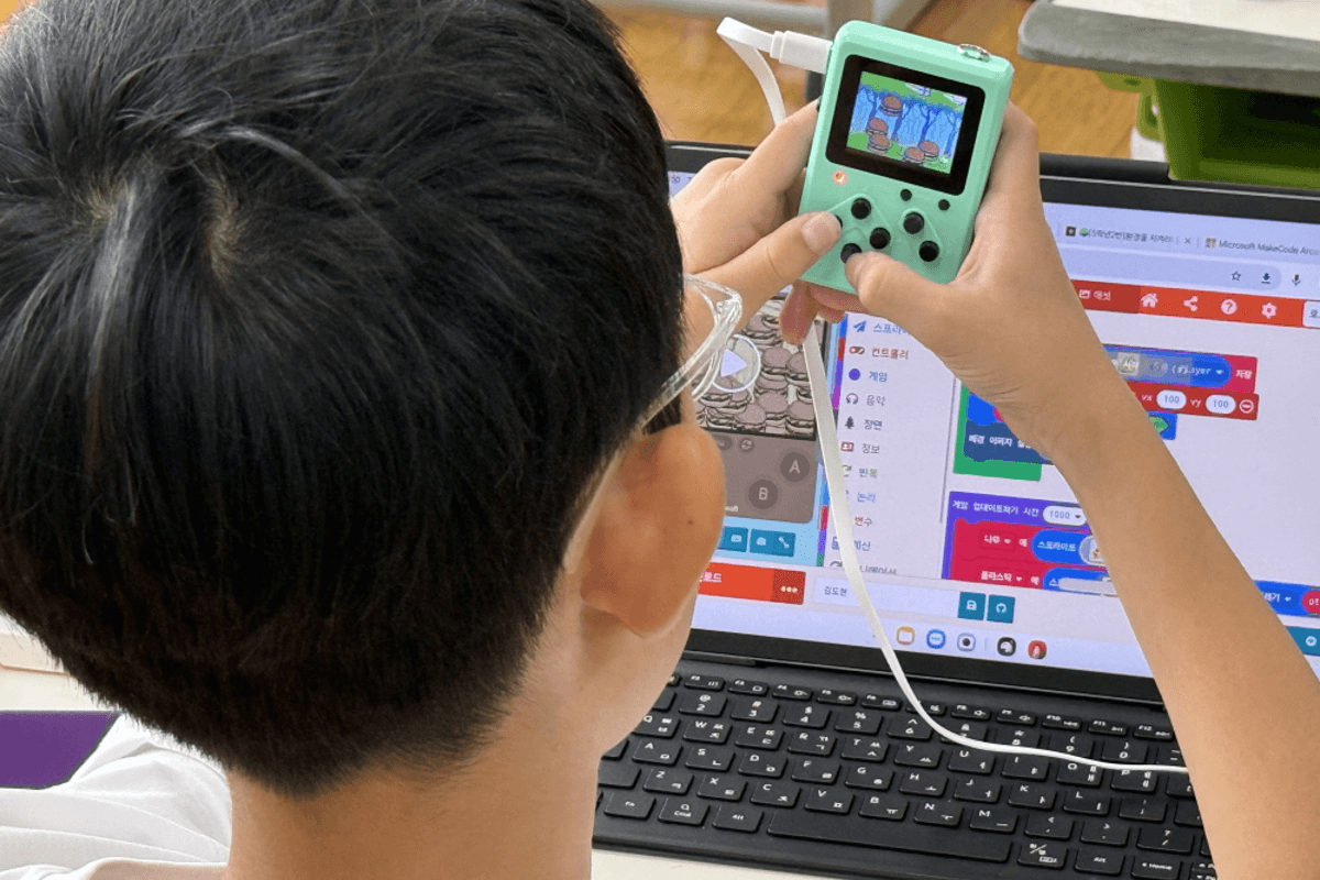 초등학교 AI·SW 수업 현장 — 휴대용 게임기 교구에서 직접 만든 게임이 실행되고 있고, 뒤편 노트북 화면에는 MakeCode Arcade 블록코딩 편집기가 떠 있다.