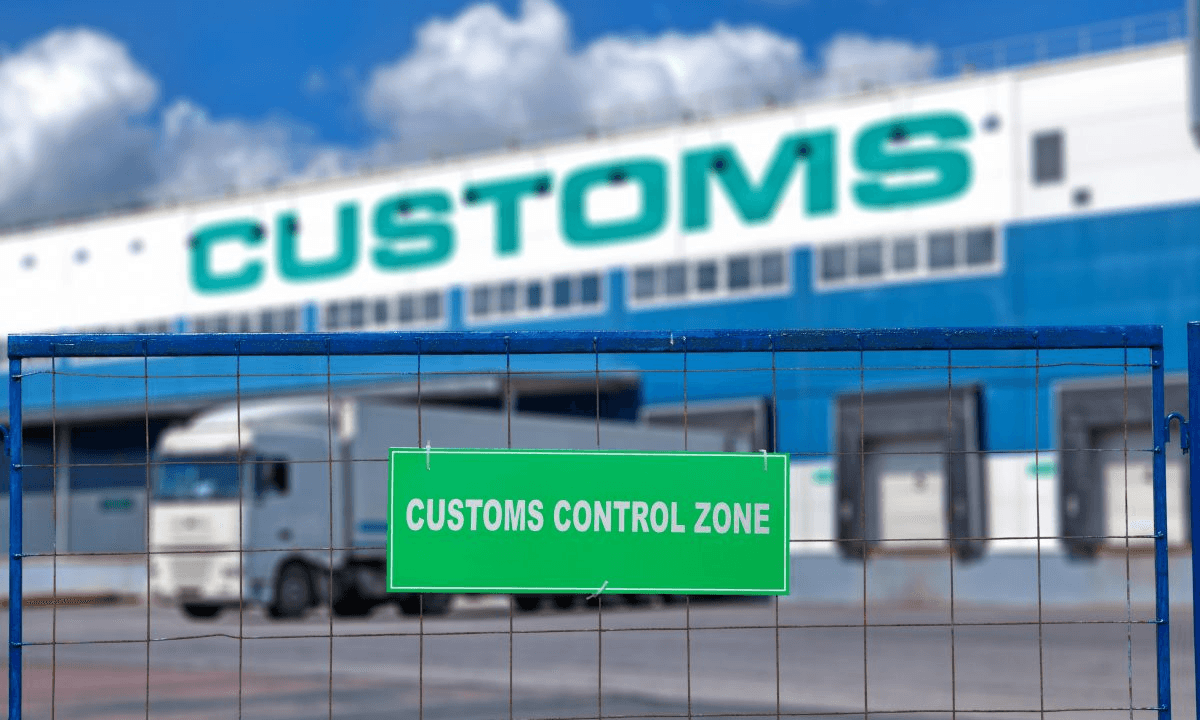 세관 통제 구역(Customs Control Zone) 표지판이 걸린 철망 너머로 화물 트럭과 세관 물류 창고가 보이는 전경