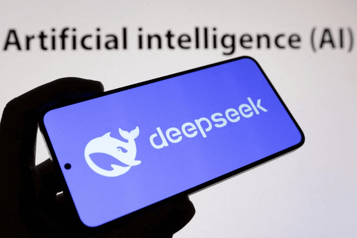 스마트폰 화면에 딥시크(DeepSeek) 고래 로고가 떠 있고 뒤편 스크린에 'Artificial intelligence (AI)' 문구가 보이는 사진 — DeepSeek의 API 가격 대폭 인상 예고를 상징한다