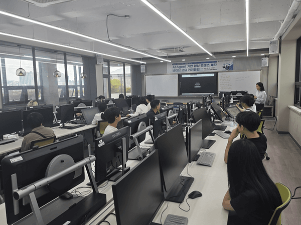 부산경상대학교 창의관 컴퓨터 실습실에서 콘텐츠 기업 재직자들이 모니터 앞에 앉아 강의를 듣고 있고, 전면 스크린에 'AI Agent 기반 영상 콘텐츠 제작 바이브 코딩 커리큘럼' 안내가 표시돼 있는 모습