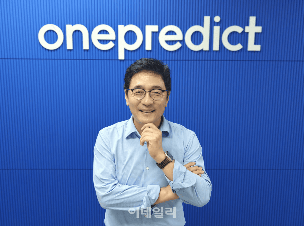 onepredict 로고가 붙은 파란색 벽 앞에서 팔짱을 낀 채 미소 짓고 있는 윤병동 원프레딕트 대표의 인터뷰 사진