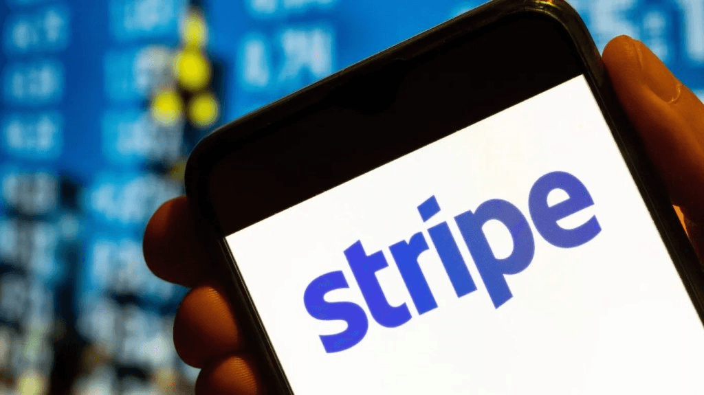스마트폰 화면에 표시된 스트라이프(stripe) 로고, 뒤로는 파란 금융 시세판이 흐릿하게 보인다