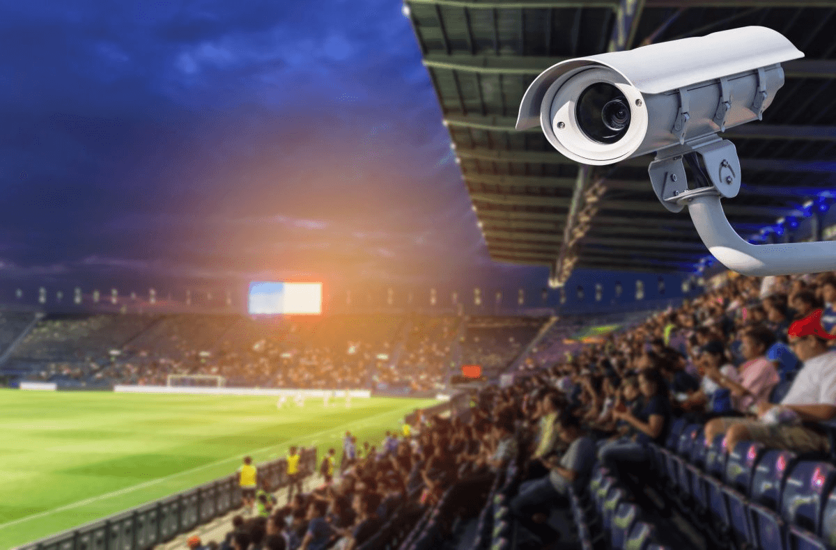 야간 축구 경기장 관중석 쪽을 향해 설치된 흰색 CCTV 감시 카메라 클로즈업. 배경에는 조명이 켜진 그라운드와 관중석이 흐릿하게 보인다.
