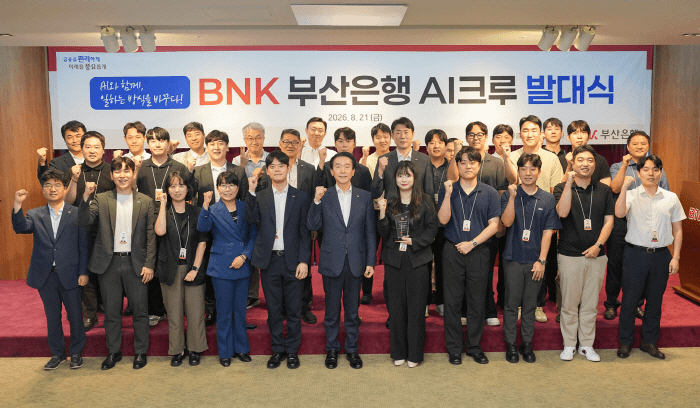 BNK부산은행 'AI크루' 발대식 단체 기념사진. 무대 배경 현수막에 'BNK 부산은행 AI크루 발대식 2026. 8. 21(금)'과 'AI와 함께, 일하는 방식을 바꾸다!' 문구, 부산은행 로고가 걸려 있고 그 앞에 사원증을 목에 건 임직원 수십 명이 주먹을 들어 보이며 서 있다.