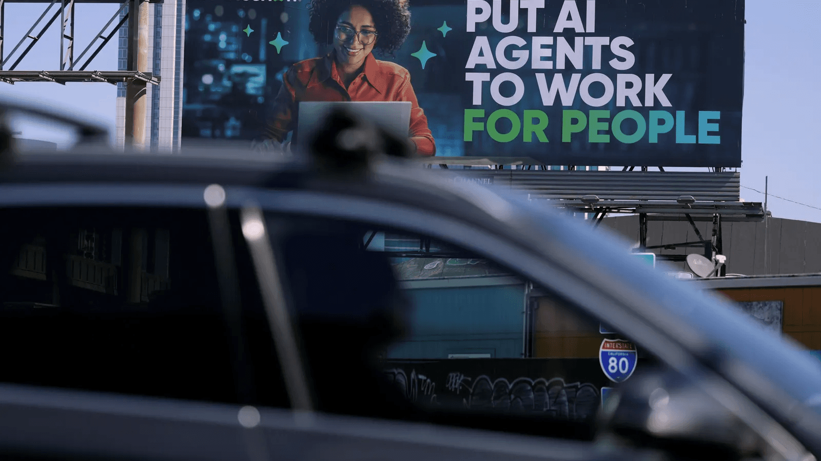 도로변 대형 전광판에 'PUT AI AGENTS TO WORK FOR PEOPLE'이라는 문구와 노트북으로 일하는 인물이 담긴 AI 에이전트 광고가 걸려 있고, 그 앞 도로를 차량이 지나가고 있다. (광고 속 인물은 기사에서 신원이 명시되지 않은 광고 모델)