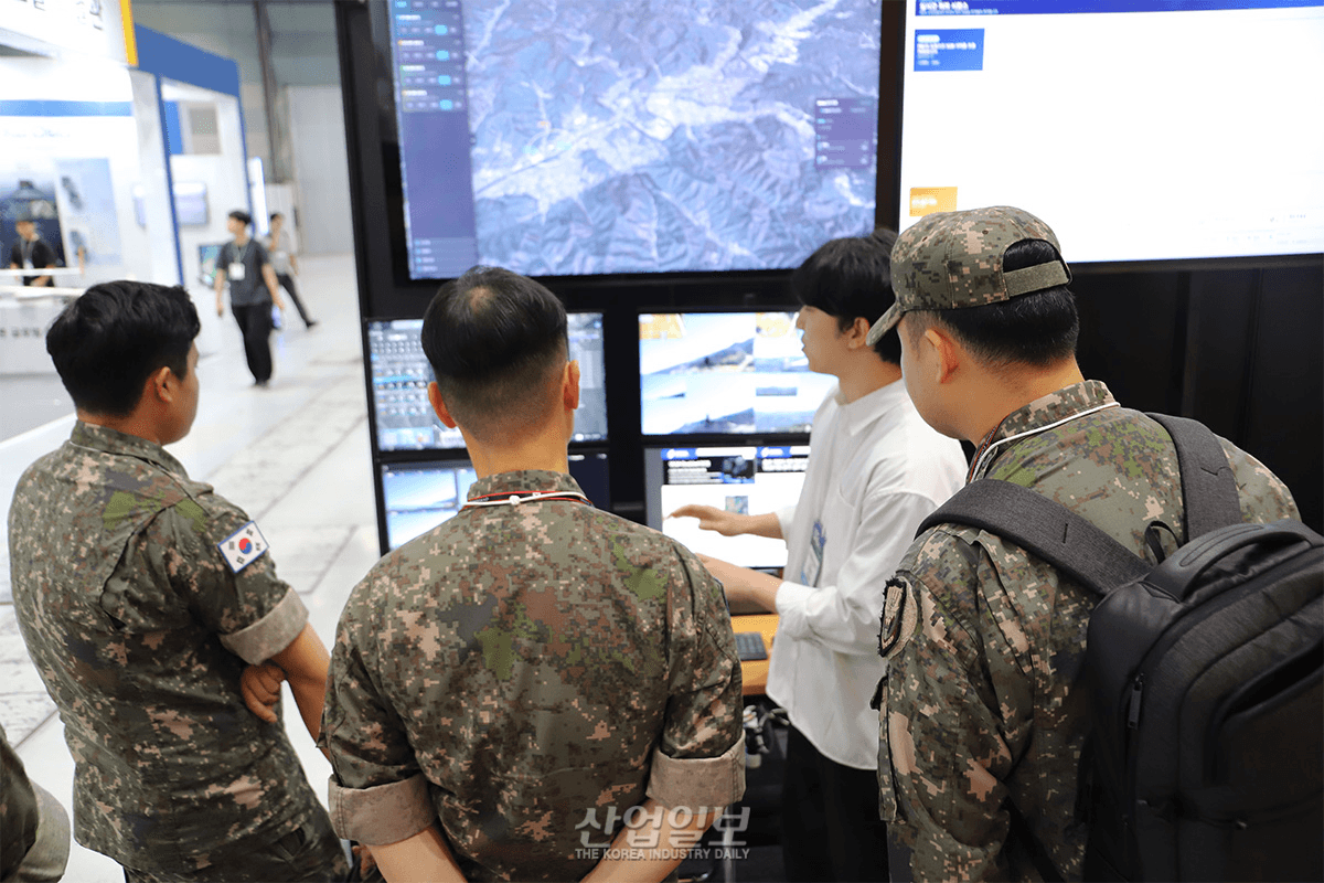 전시 부스에서 군복을 입은 관람객들이 무인기 관제 화면(지형 지도와 실시간 영상 패널)을 보며 담당자의 시연 설명을 듣고 있다