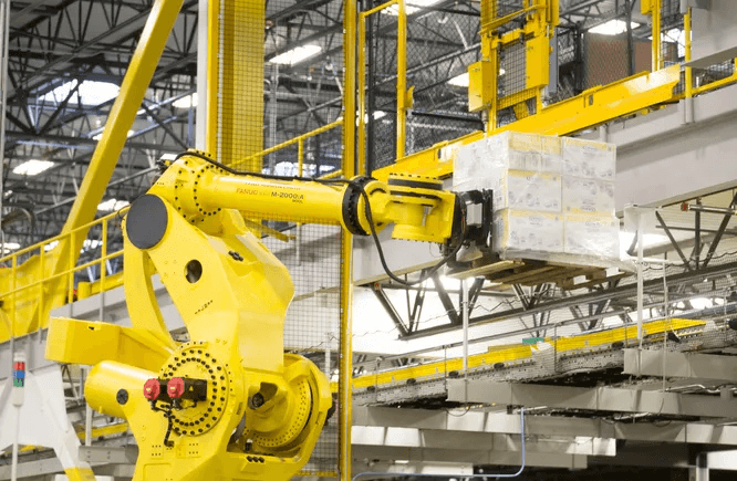 공장 물류 라인에서 노란색 대형 산업용 로봇(FANUC M-2000iA)이 적재 팔레트를 들어 올리는 모습